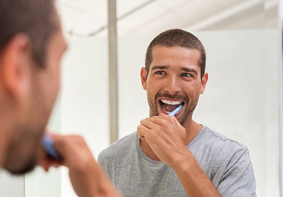 A smiling man brushing his teeth in front of a bathroom mirror